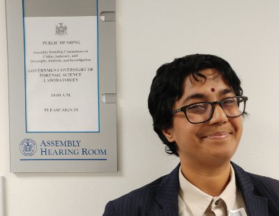 Smiling Sumana, wearing a collared shirt and jacket, next to an "Assembly Hearing Room" sign-holder with the paper agenda for the public hearing mentioned in the post
