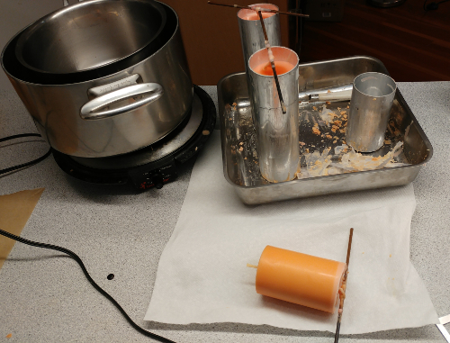 Candlemaking items on countertop: pots on a burner, molds in tray, and an orange candle on a napkin, wick still tied to stabilization stick.