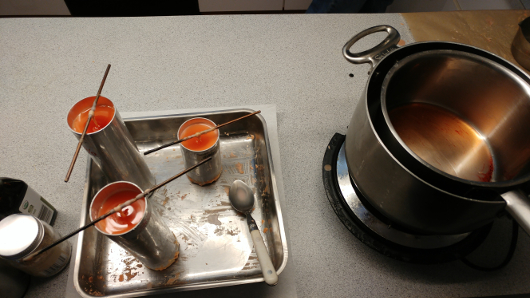 Candlemaking items on countertop: three cylindrical metal molds filled with hot reddish wax, a wax-encrusted tray and spoon, and molten wax in pots atop a burner.