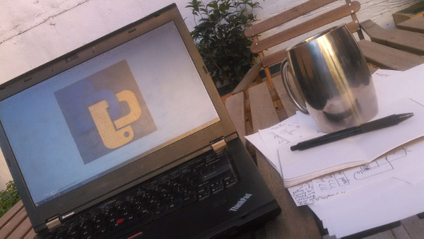 Laptop displaying bpython logo next to half-scratched-out bpython logo, Python code, and technical prose written and drawn on paper, with notebook and pen and mug, on a wooden table